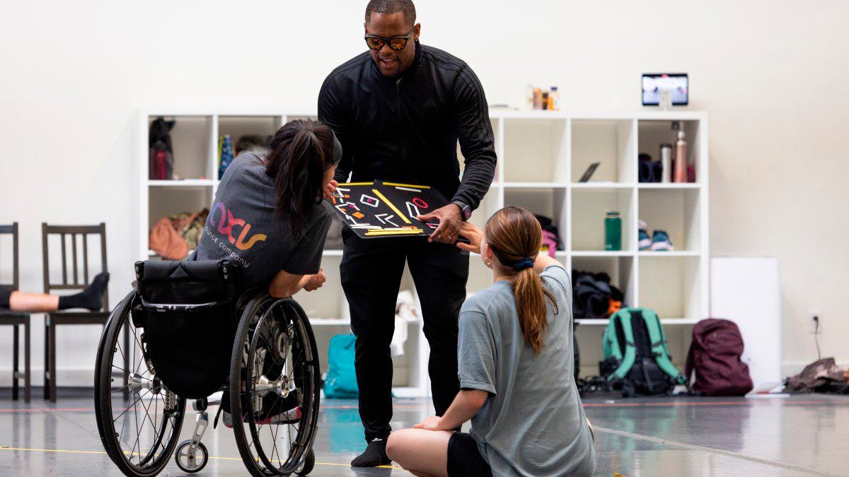 AXIS Choreo-Lab Fellow DJ Robinson, who is blind, shares a tactile map of a dance studio with AXIS dancers Julie Hasushi & Anna Gichan. Julie sits in her wheelchair, Anna sits cross-legged on the floor.